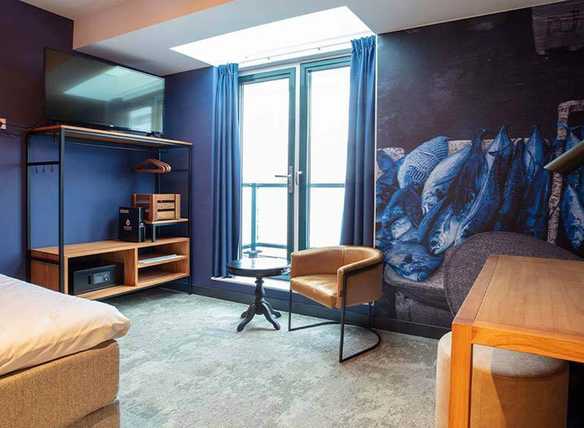 A hotel room with a bed, a chair, a small table, a TV, and a mural.