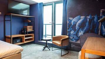 A hotel room with a bed, a chair, a small table, a TV, and a mural.