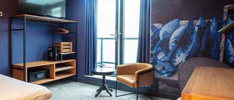 A hotel room with a bed, a chair, a small table, a TV, and a mural.