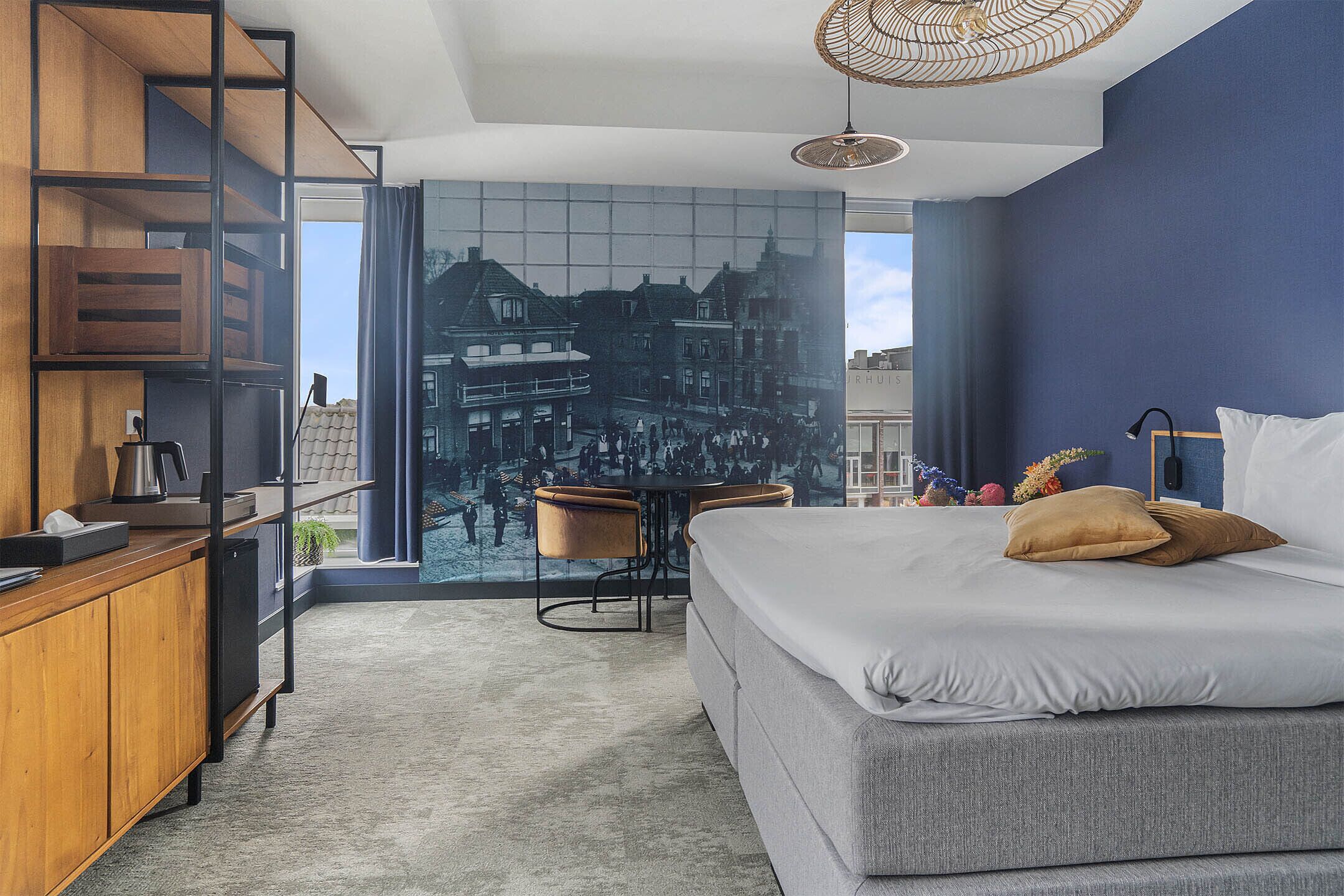 A modern hotel room with a large bed, a desk with a chair, and a view of a cityscape through a large window.