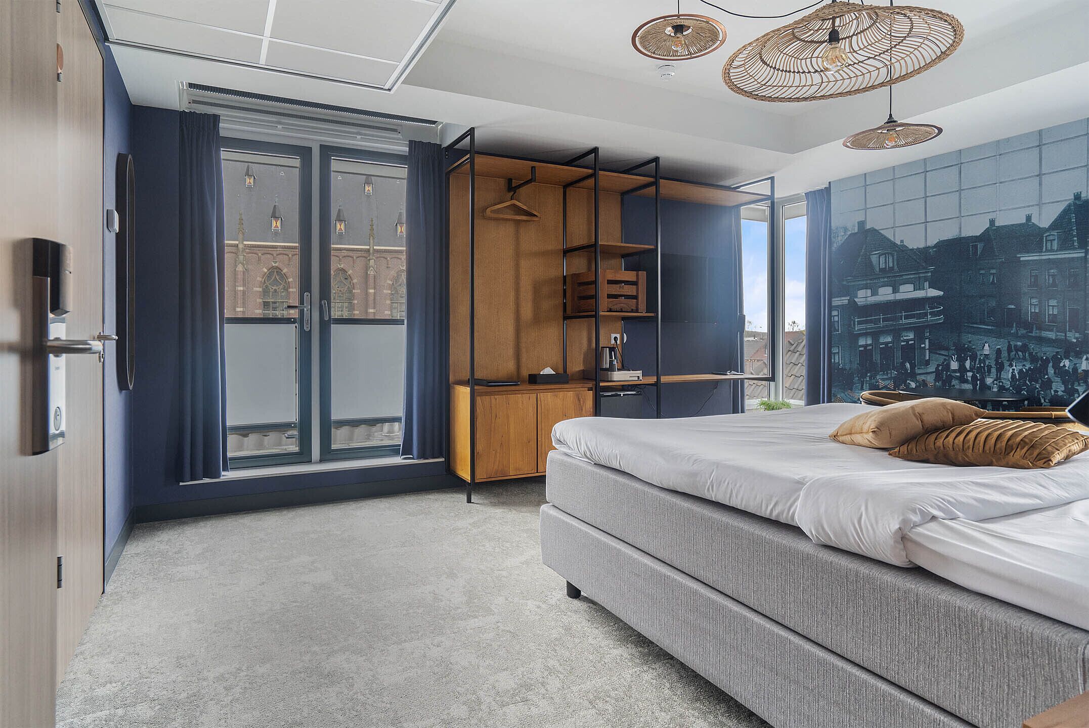 A modern bedroom with a large bed, a wardrobe, a desk, and a view of a cityscape through a glass door.