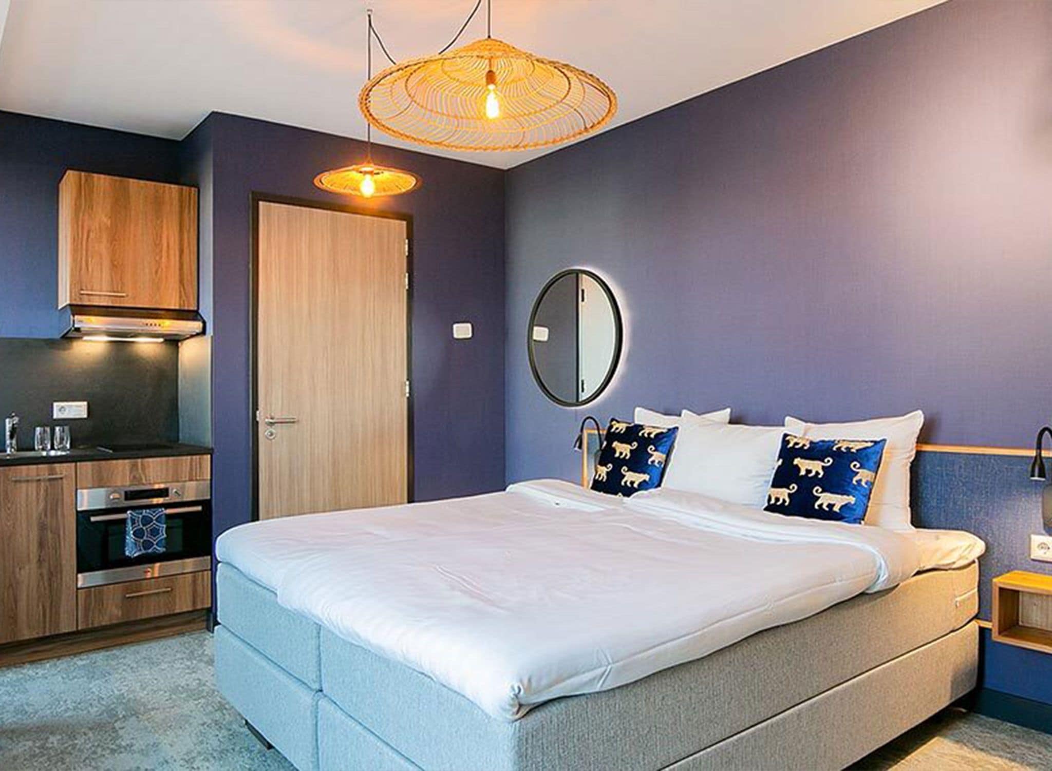 A bedroom with a bed, a bedside table, a mirror, a wardrobe, and a kitchenette area.