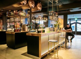 Zona de bar con un cantinero, estantes llenos de botellas de licor, vasos y una máquina de café.