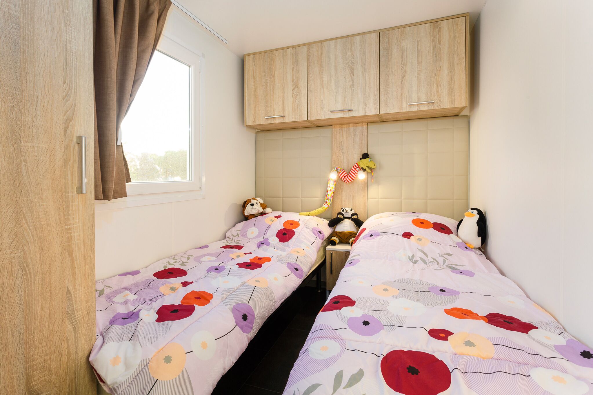 A bedroom with two beds, wooden cabinets, a window with curtains, and stuffed toys on the beds.