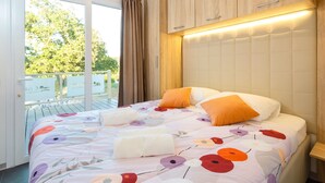 Une chambre à coucher avec un lit, une tête de lit en bois, une fenêtre avec des rideaux et une vue sur la verdure à l’extérieur.