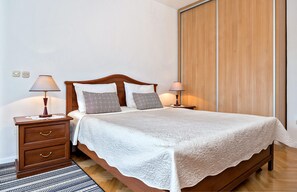 A neatly made bed with a white quilt, two bedside lamps, and a wooden nightstand with drawers. - Apartments Emi (Split)