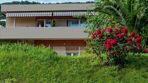 Un edificio de dos pisos con techo plano, un balcón con barandilla y un gran arbusto de flores rojas en la fachada.