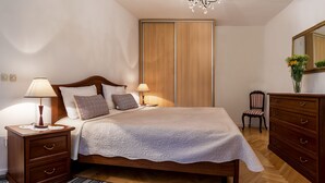 Una cama bien tendida con colcha blanca, dos lámparas de noche y almohadas estampadas. Se ve un aparador de madera con cajones y una silla con un florero.