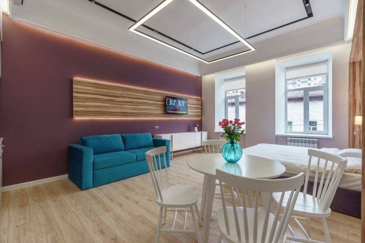 Habitación de hotel moderna con sofá color turquesa, mesa de comedor blanca y sillas, televisor montado en la pared y ventanas grandes que ofrecen vistas al exterior.
