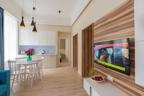 A modern living room with a flat-screen TV mounted on a wooden wall, a dining area with white chairs and a table, and a kitchen in the background.