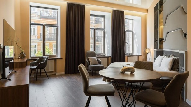 Une chambre d’hôtel moderne avec une table à manger ronde, deux fauteuils, un canapé et un téléviseur. De grandes fenêtres, équipées de rideaux, laissent entrer la lumière naturelle et offrent une vue sur la ville.