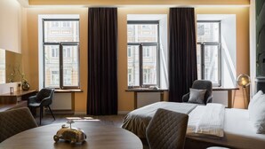 Une chambre d’hôtel avec un lit, une table ronde sur laquelle est posée une voiture jouet, deux chaises, un bureau avec une lampe et de grandes fenêtres avec des rideaux.