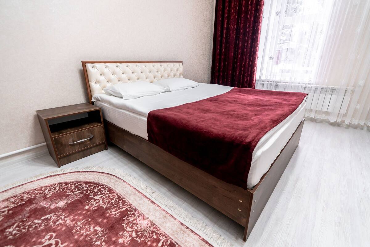 A neatly made bed with a red blanket, a wooden nightstand with a drawer, and a patterned rug on a light-colored floor.