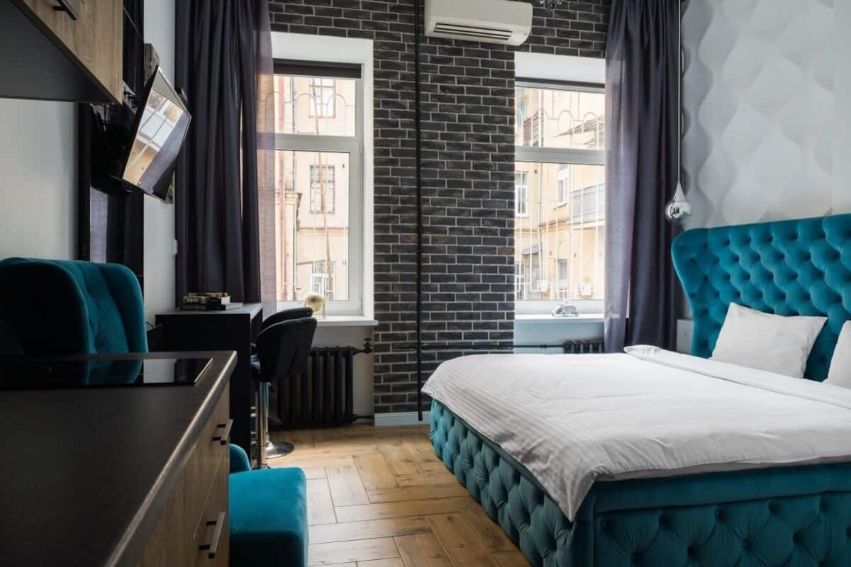 A hotel room with a large bed, a desk, and a chair. There are windows with curtains, a radiator, and a wall-mounted TV.