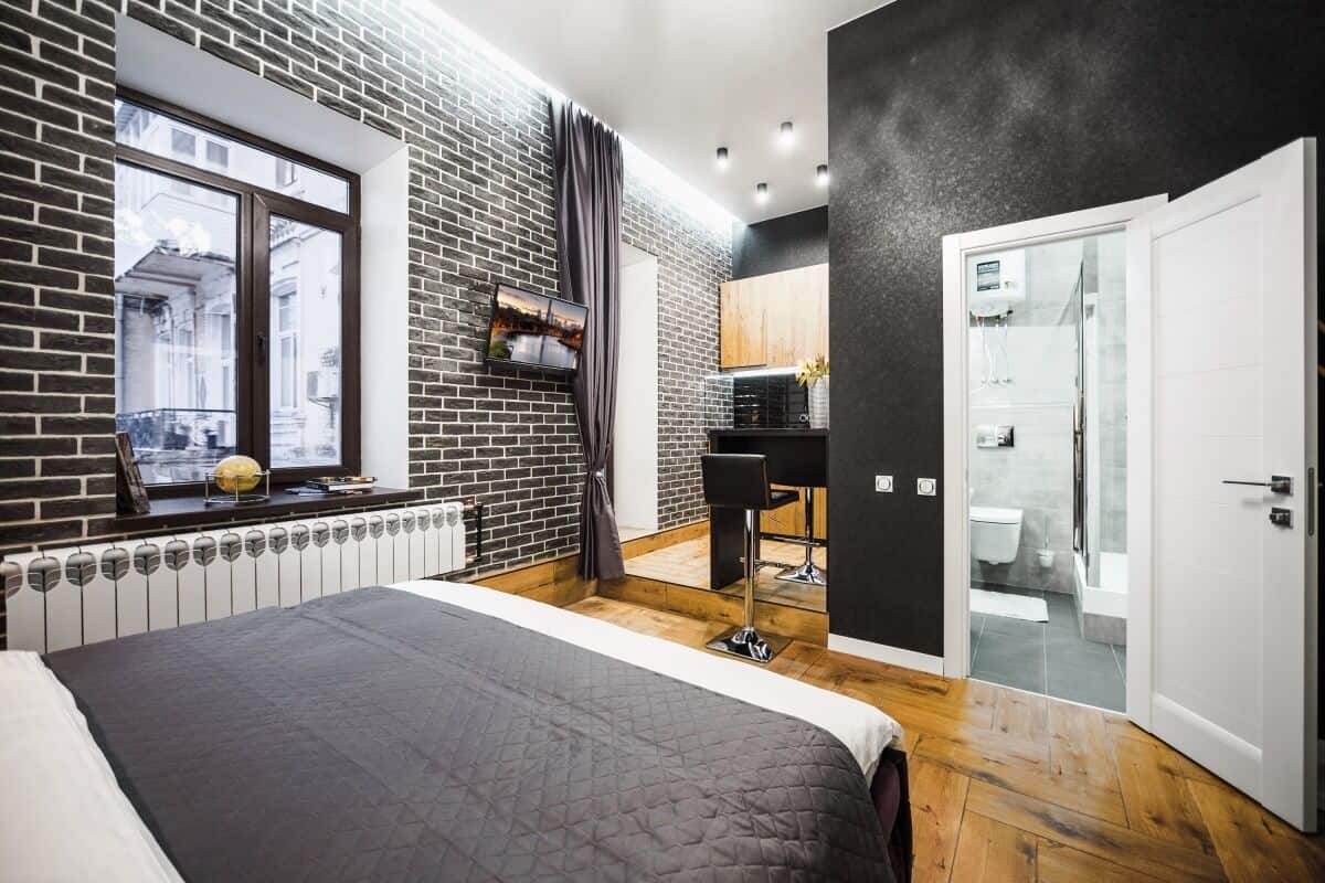 A modern hotel room with a brick wall, a large bed, a small desk, and a bathroom visible through a glass door.
