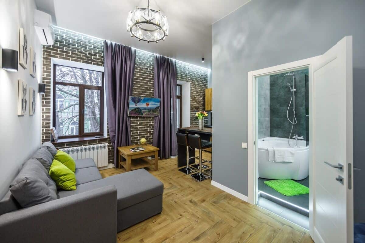 A modern living room with a grey sofa, a wooden coffee table, and a TV on a brick wall. There's a glass-enclosed shower area with a white bathtub.