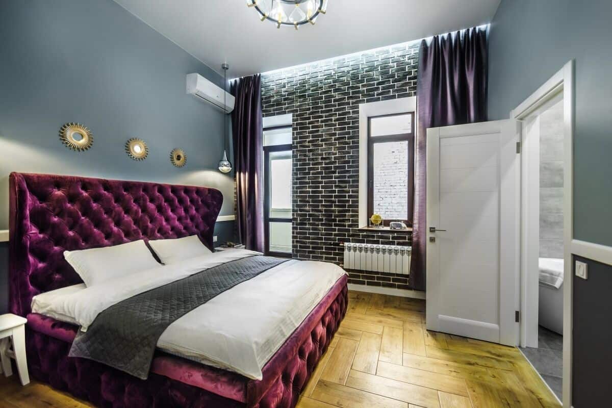 A bedroom with a tufted headboard, a bed with white and gray bedding, a brick-patterned wall, a window with curtains, and a white door.