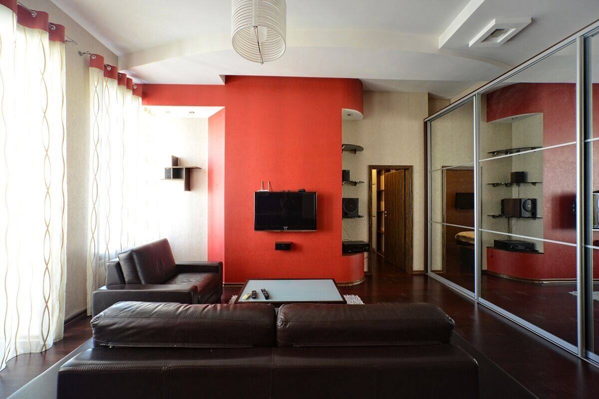 A modern living room with a red accent wall, a flat-screen TV, leather sofas, a coffee table, and a glass partition with shelves.