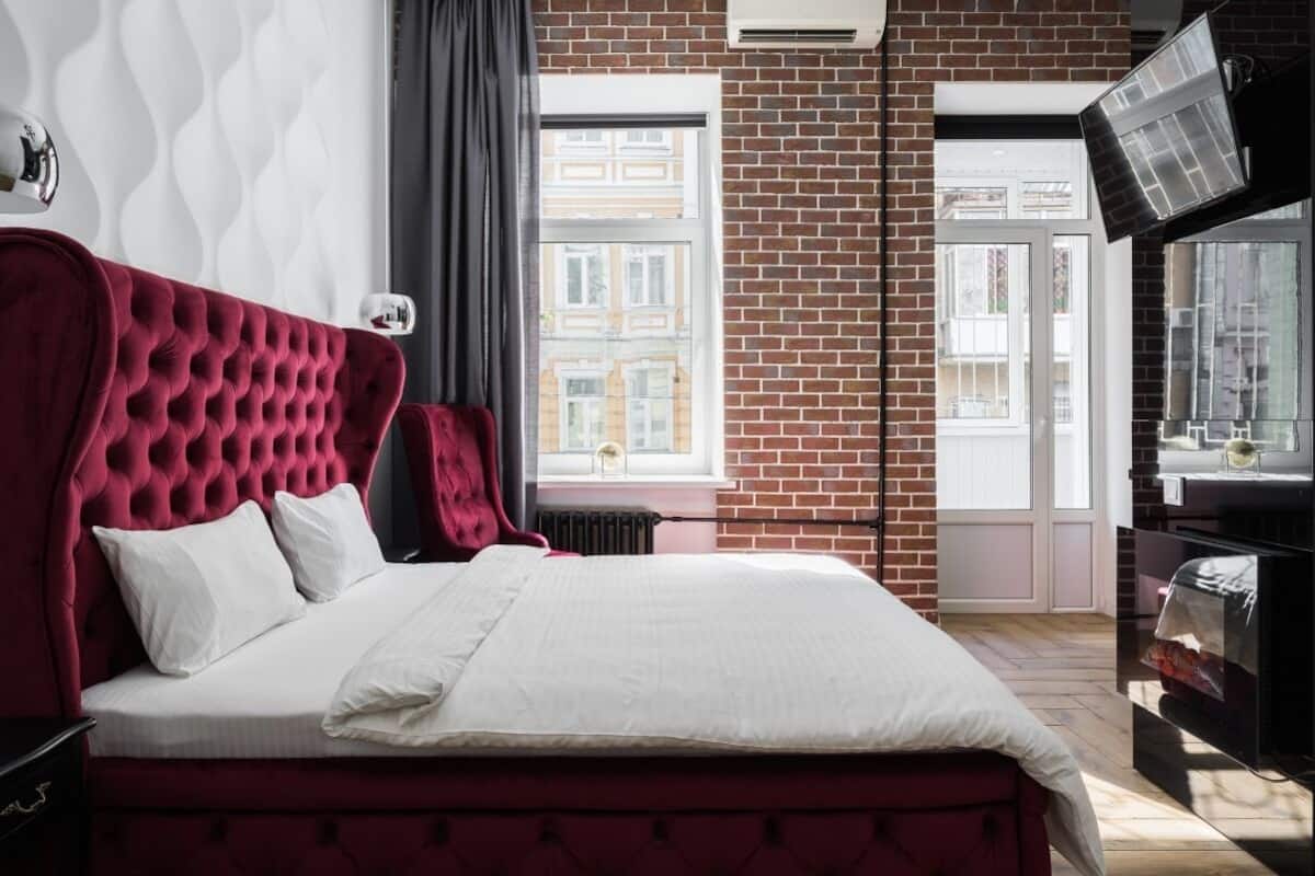 A bedroom with a red upholstered headboard, white bedding, and a brick wall.