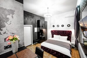 A modern bedroom with a large bed, a desk, and a chair. There is a dresser, a mirror, and a chandelier.