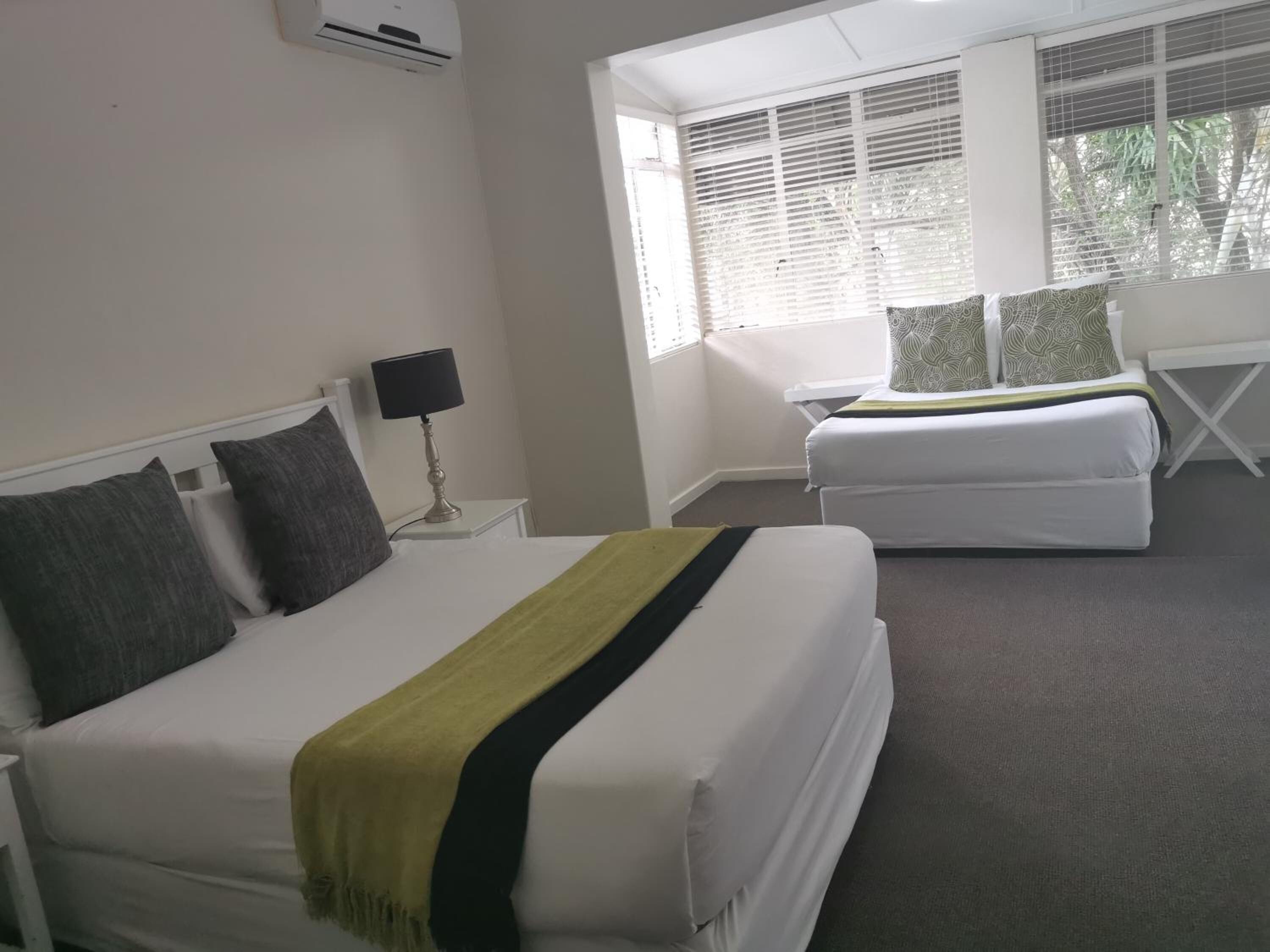 A hotel room with two beds, a bedside table with a lamp, and a window with blinds.