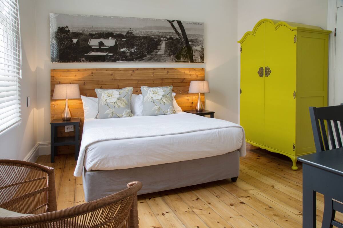 A bedroom with a bed, two bedside tables with lamps, a wicker chair, a wooden floor, and a bright yellow wardrobe.