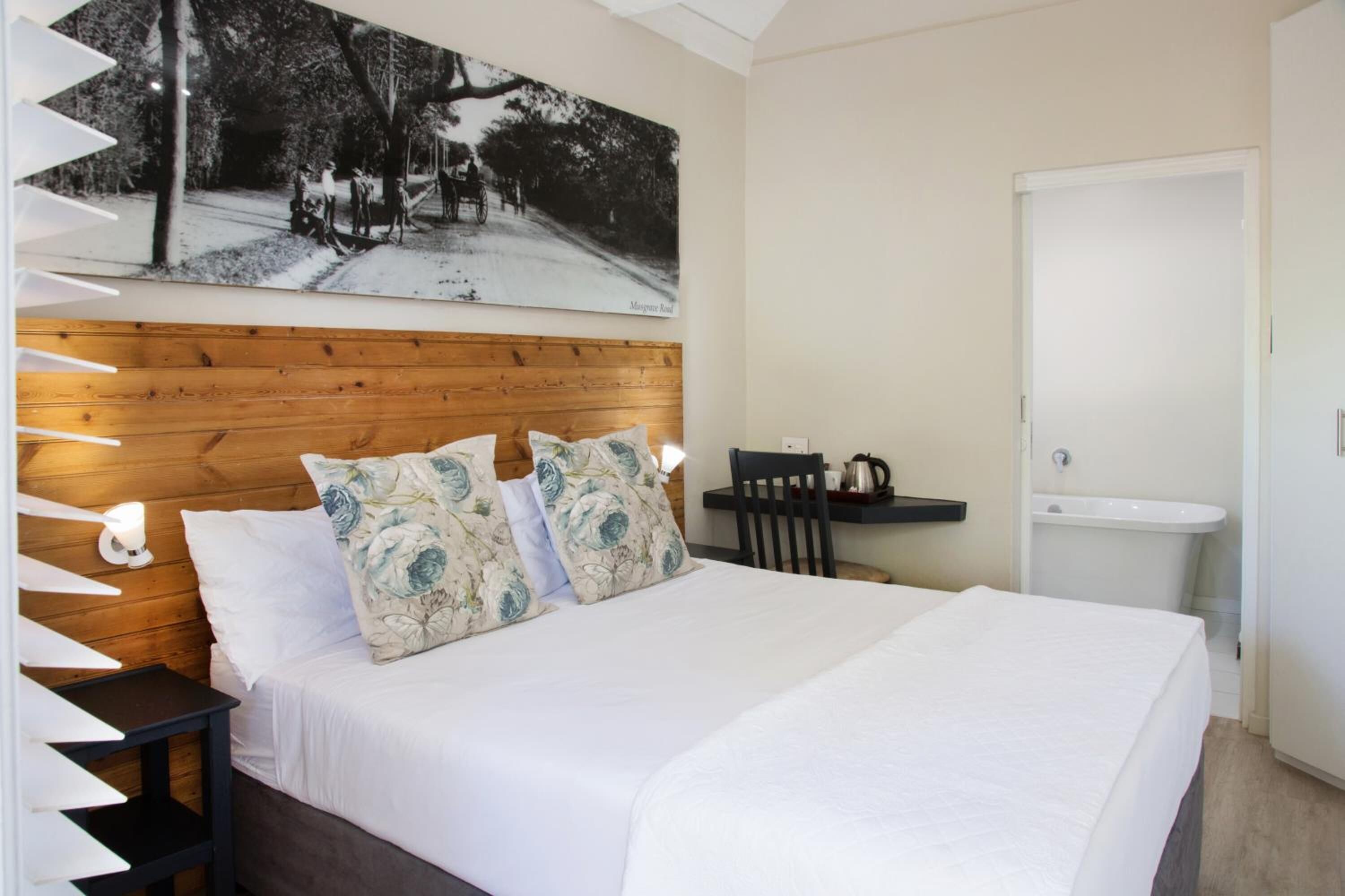 A bedroom with a wooden headboard, a bed with white linens, a nightstand with a lamp, a painting on the wall, a small table with a kettle, and a white sink.