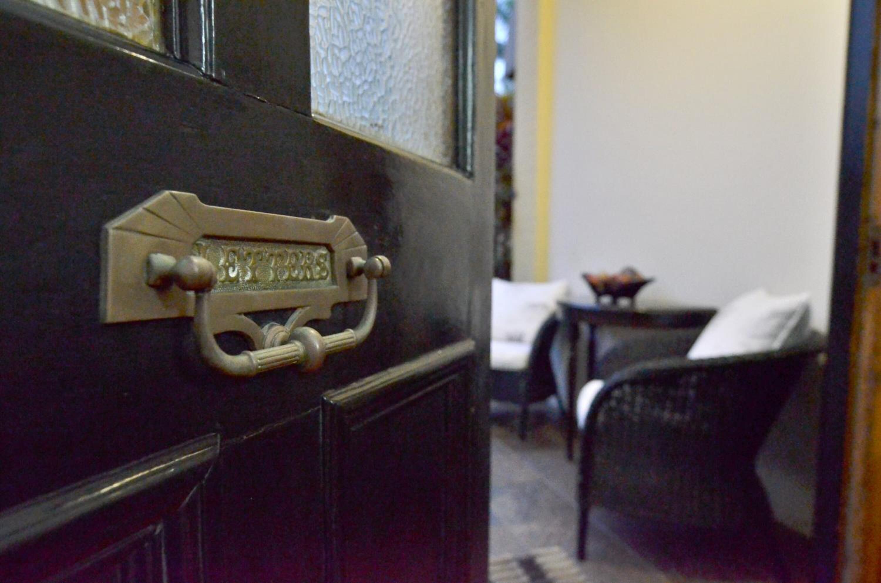Une porte dotée d’une poignée en laiton et d’une plaque nominative, une chaise noire et une petite table surmontée d’un coussin blanc.
