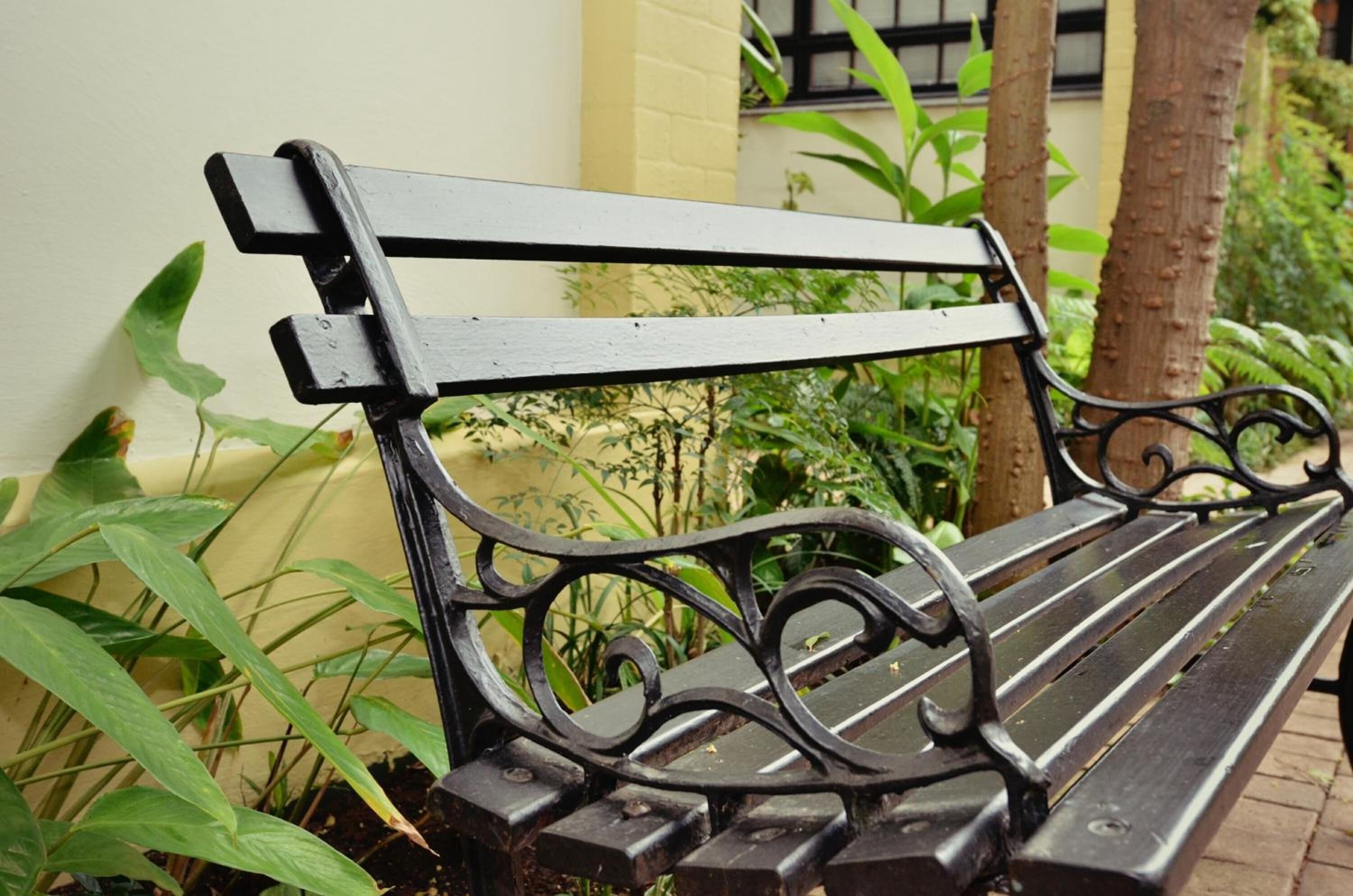Un banc en métal noir orné de motifs travaillés, placé à l’extérieur, avec de la verdure et un bâtiment en arrière-plan.