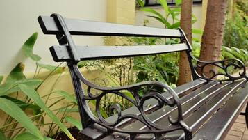 Un banc en métal noir orné de motifs travaillés, placé à l’extérieur, avec de la verdure et un bâtiment en arrière-plan.