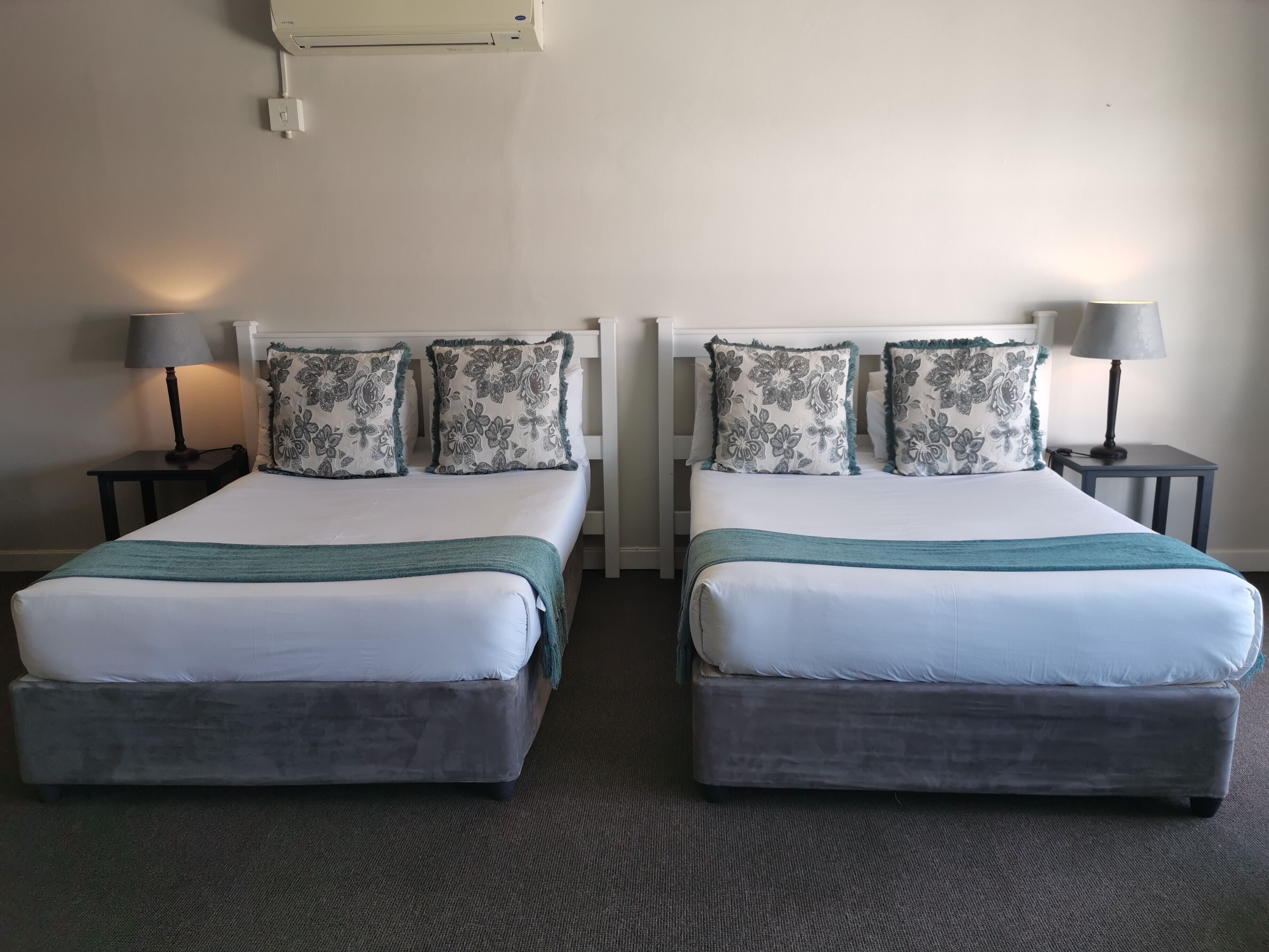 Quarto de hotel com duas camas, cada uma com colchão branco e cabeceira cinzenta. Há duas mesas de cabeceira com abajures, e o chão é coberto por carpete.