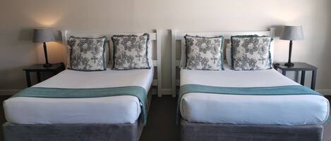 Quarto de hotel com duas camas, cada uma com colchão branco e cabeceira cinzenta. Há duas mesas de cabeceira com abajures, e o chão é coberto por carpete.