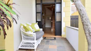 Un estrecho pasillo exterior con una silla de mimbre blanca y cojines, suelo de baldosas y una escalera de madera que sube a un nivel superior.