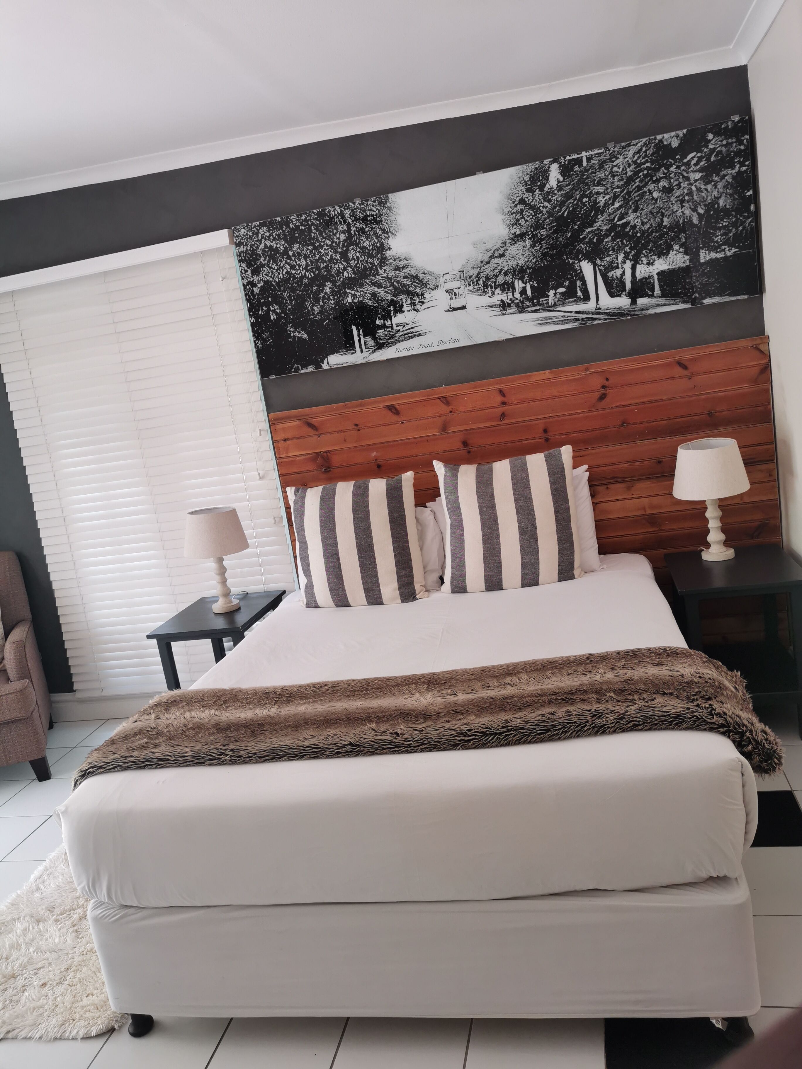 Un dormitorio con cama, dos mesitas de noche, cabecera de madera, una fotografía grande en blanco y negro y una ventana con persianas.