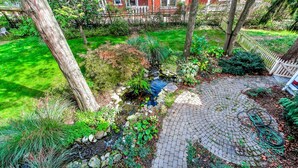 Un jardín con un camino de ladrillo circular, un pequeño elemento acuático y una cerca blanca de piquetes.