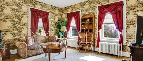 A living room with floral wallpaper, a sofa, a coffee table, a bookshelf, and red curtains.