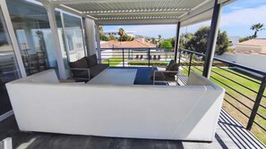 A covered outdoor seating area with a white sofa and a small table, overlooking a residential area with houses and a clear sky. - Detached villa private pool and sea views (Sotogrande)