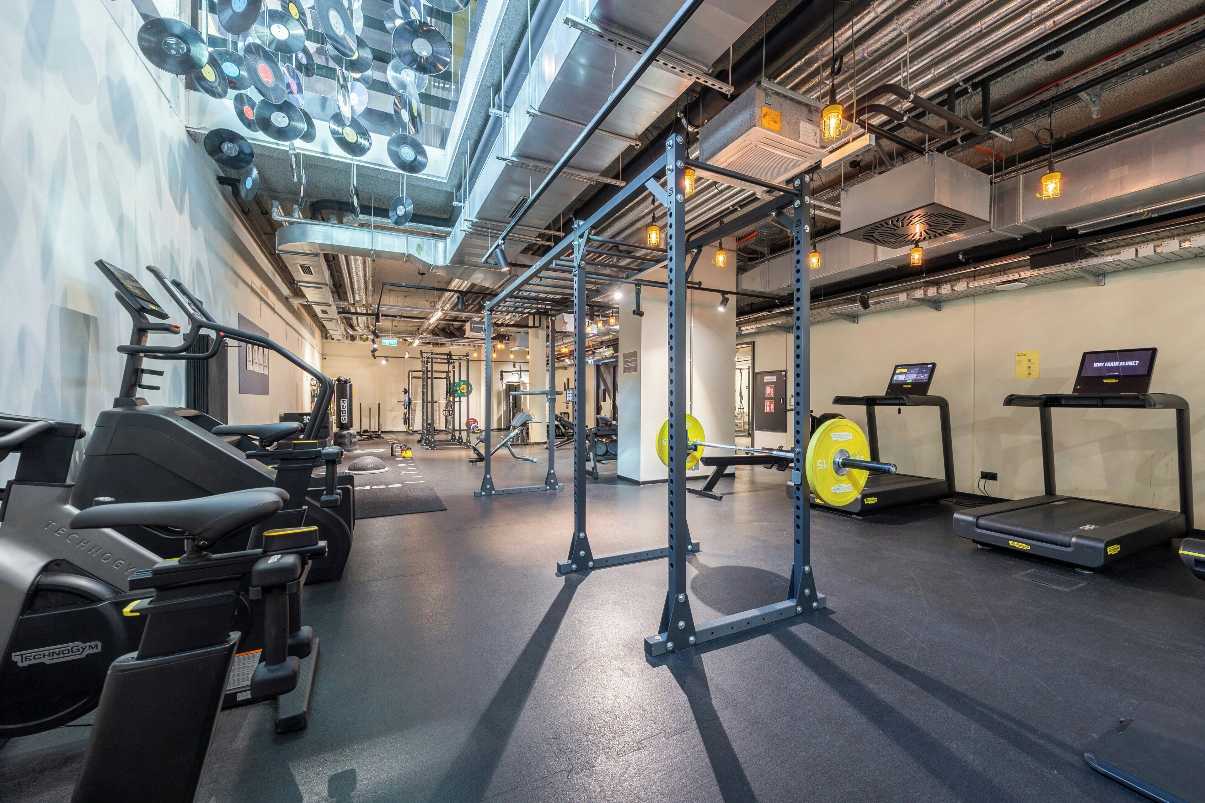 Een fitnessruimte met trainingsapparatuur, een wandrek en een digitaal scherm.