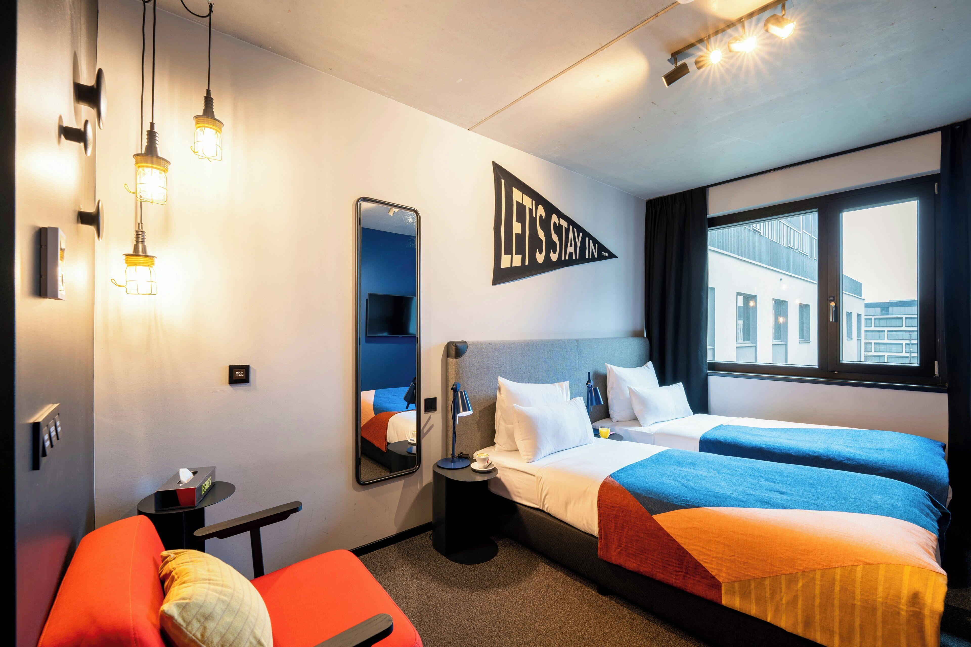 Hotellihuone, jossa on kaksi sänkyä, punainen sohva, pieni pöytä, yöpöytä, peili ja ikkuna, josta on näkymä rakennuksiin.