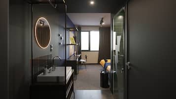 Habitación de hotel moderna con cabina de ducha de vidrio, un escritorio con silla, cama con manta azul y un amplio ventanal que proporciona luz natural.