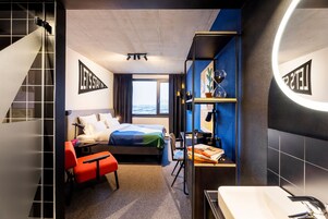 Quarto de hotel com cama, escrivaninha, cadeira, estante de livros, espelho e janela com vista.