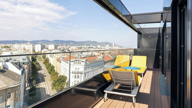 Uma varanda com vista para a paisagem urbana, incluindo edifícios e uma cadeia montanhosa ao longe.