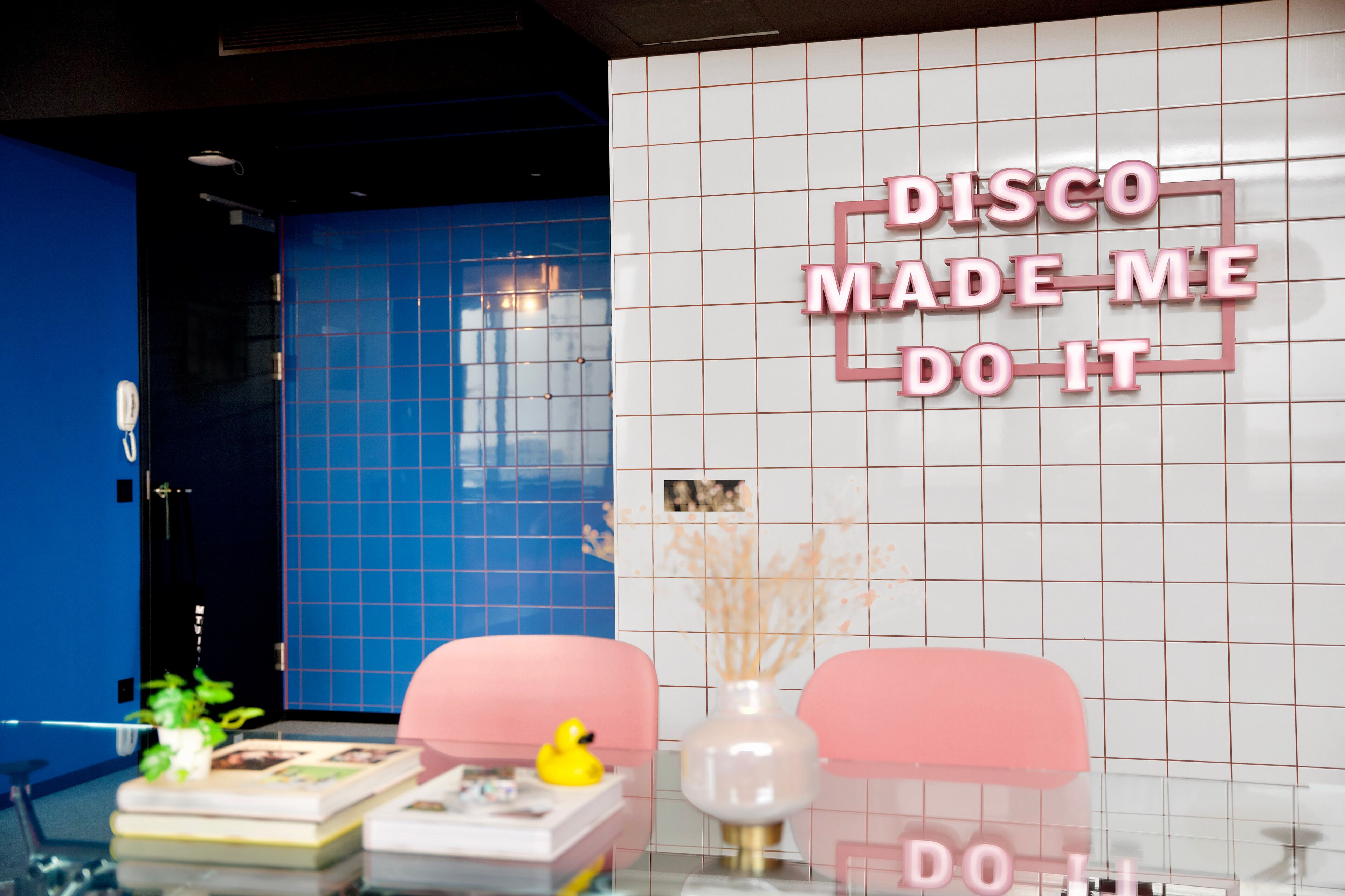 A room with a blue wall, a white tiled wall with a neon sign, a pink chair, a yellow rubber duck, and a vase with dried plants.