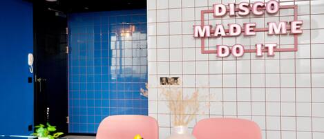A room with a blue wall, a white tiled wall with a neon sign, a pink chair, a yellow rubber duck, and a vase with dried plants.
