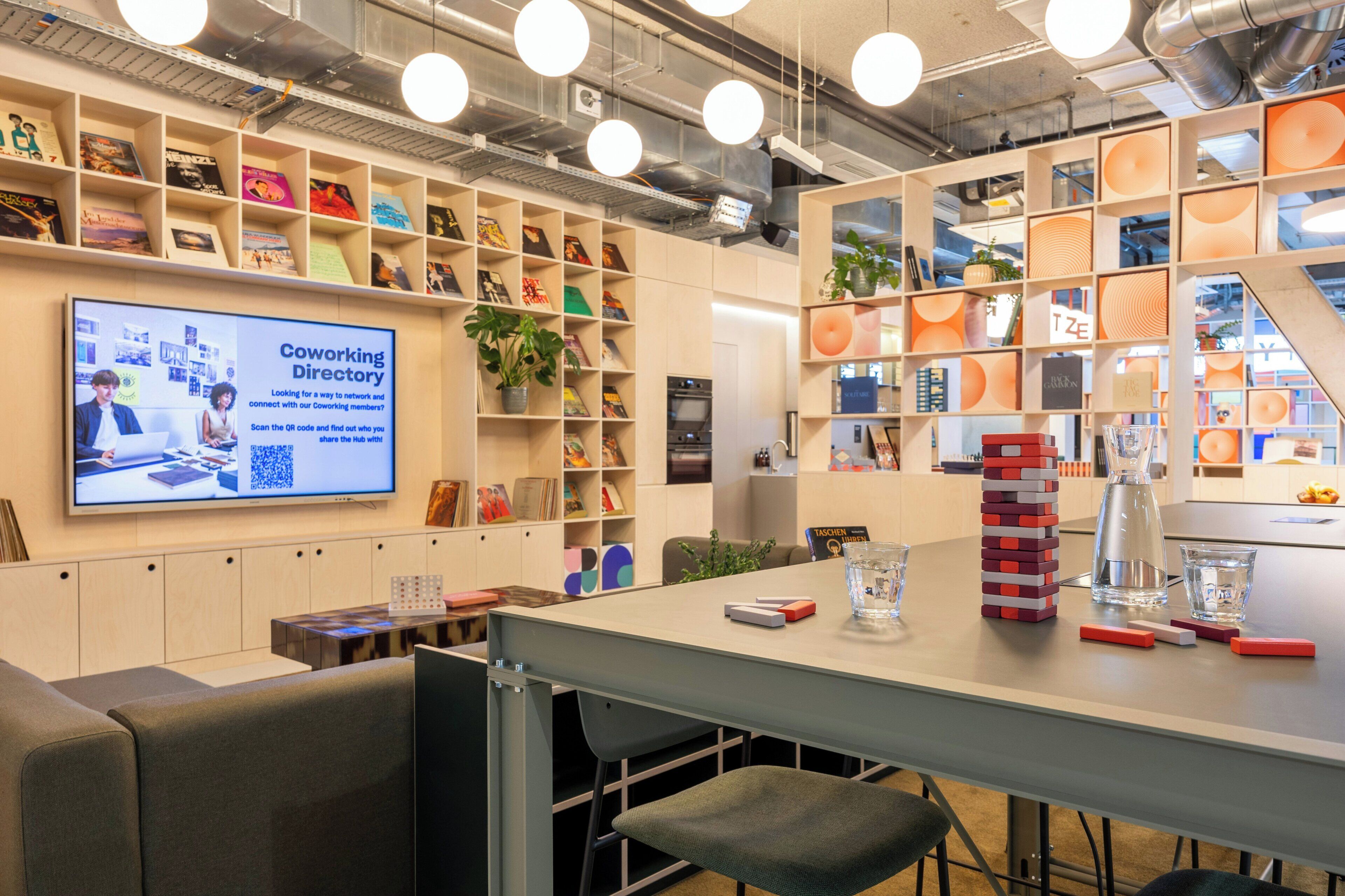 A coworking space with a large screen displaying a presentation, shelves with books and plants, and a table with a stack of books and glasses.