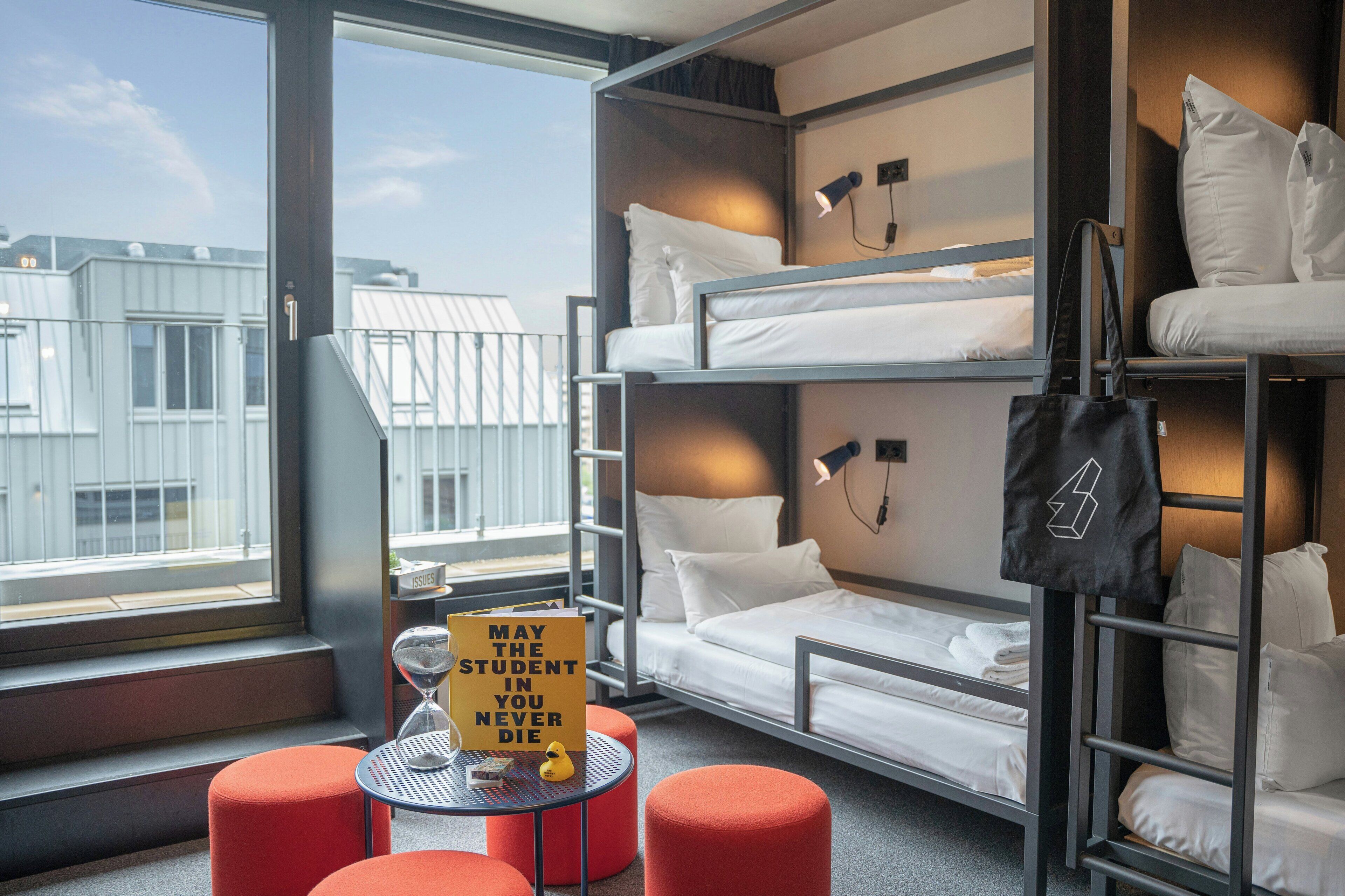 A bunk bed room with a table and ottomans, a window with a view of buildings, and a sign on the table.