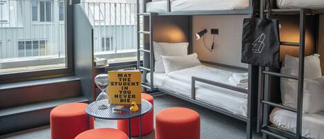 A bunk bed room with a table and ottomans, a window with a view of buildings, and a sign on the table.