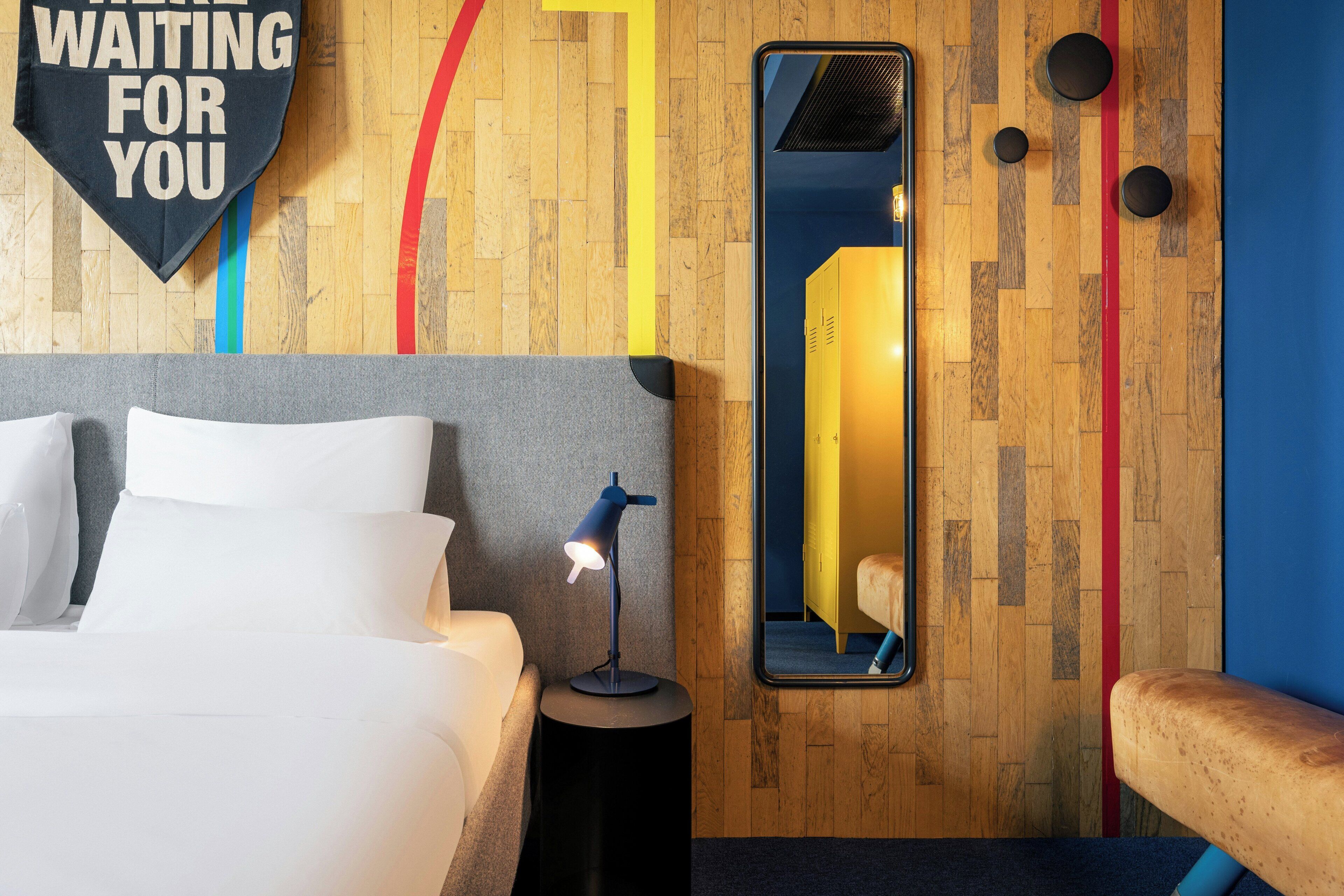 Ein Hotelzimmer mit Bett, Nachttisch mit Lampe, Spiegel und einem Schild mit der Aufschrift 'WAITING FOR YOU'.