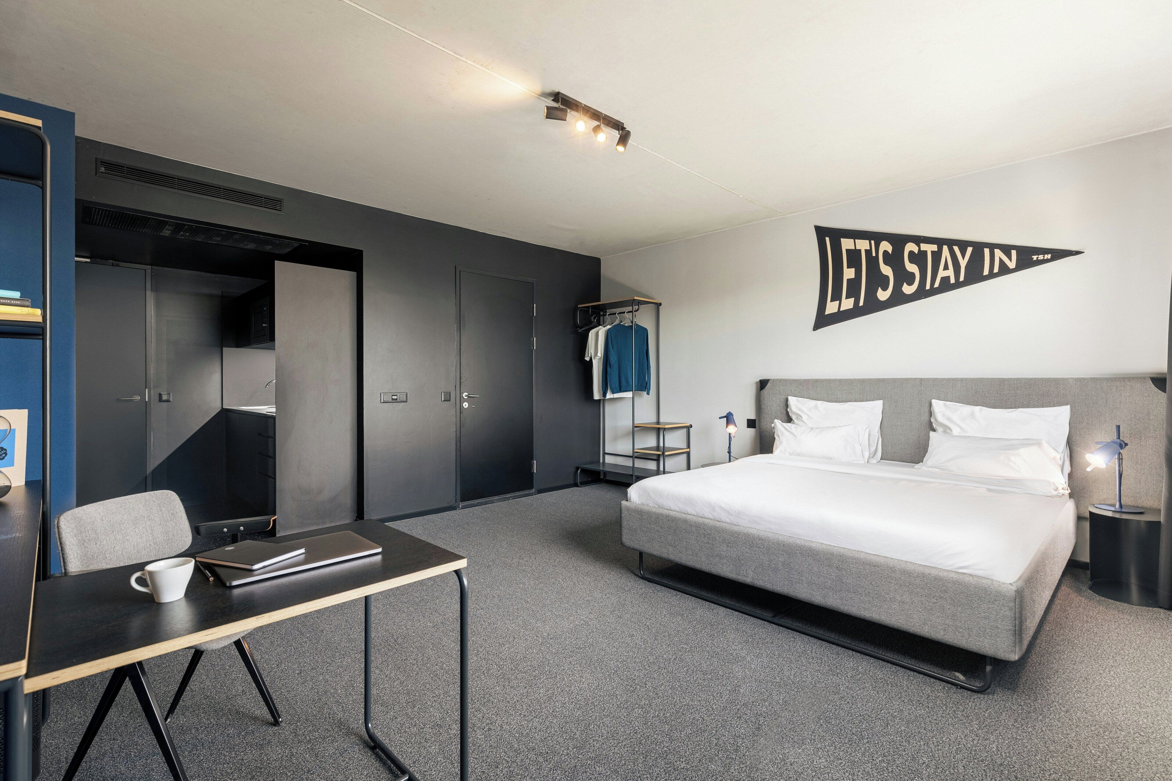 A modern hotel room with a bed, a desk, a chair, a wardrobe, and a wall-mounted sign.