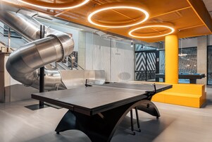 Uma mesa de pingue-pongue num espaço interior moderno, com estrutura metálica e detalhes em amarelo.